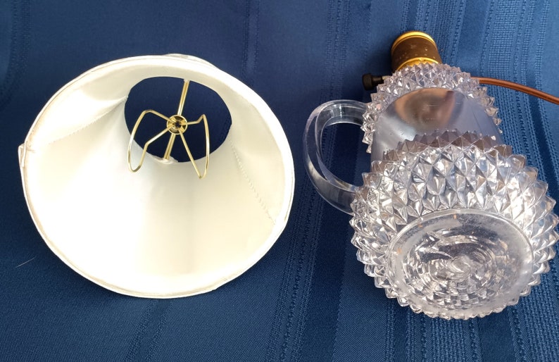 May include: A white lampshade with a gold metal harp and a clear glass lamp base with a diamond pattern.