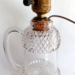 May include: A clear glass lamp with a diamond-shaped pattern and a handle. The lamp has a brass socket with a black switch and a brown cord.
