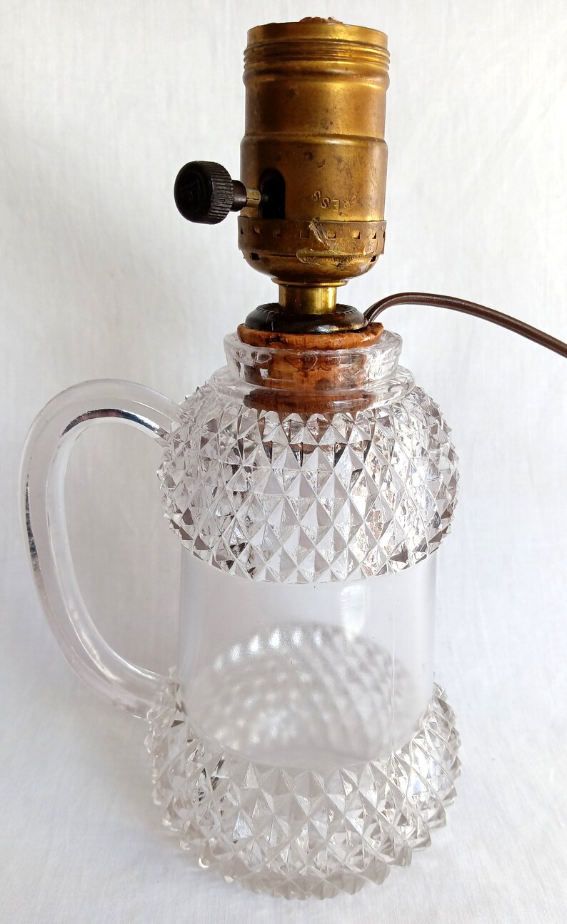 May include: A clear glass lamp with a diamond-shaped pattern and a handle. The lamp has a brass socket with a black switch and a brown cord.