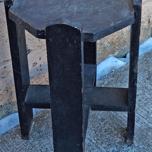 May include: A small, black, wooden side table with a hexagonal top and a lower shelf. The table has four legs and is constructed with simple joinery. The finish is worn, giving it a vintage appearance. The table is approximately 24 inches tall.