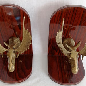 May include: A pair of bookends made of brass moose heads mounted on wood bases with a dark red wood grain finish.