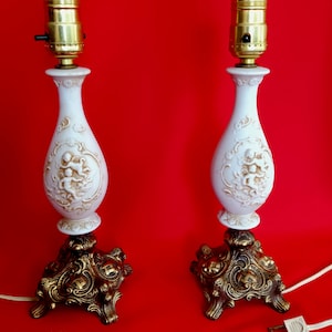 May include: A pair of white ceramic table lamps with gold accents and cherub designs. The lamps have a classic, vintage style and are perfect for adding a touch of elegance to any room.