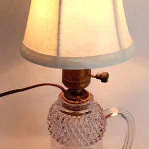 May include: A clear glass lamp base with a diamond pattern and a handle. The lamp has a white fabric lampshade with a floral pattern.