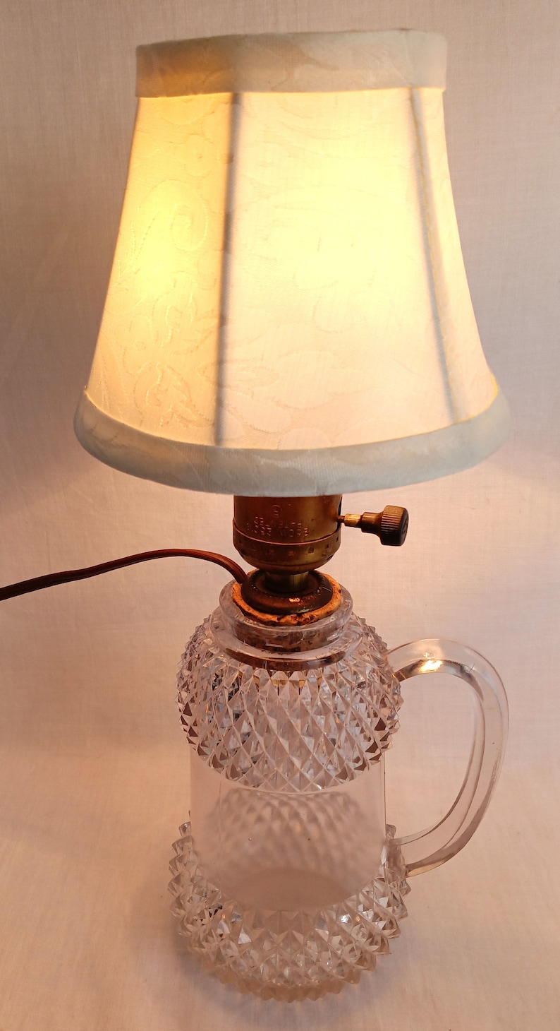 May include: A clear glass lamp base with a diamond pattern and a handle. The lamp has a white fabric lampshade with a floral pattern.