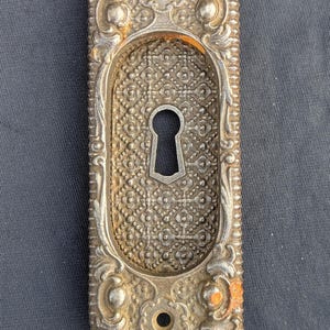 May include: Ornate, antique silver-toned door plate with a keyhole. The rectangular plate features an oval keyhole and is surrounded by detailed floral and scrollwork. Two holes are visible at the top and bottom. The metal shows signs of age and wear.