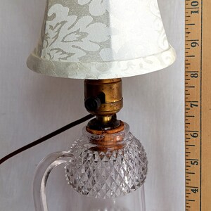 May include: A vintage glass lamp base with a clear, faceted design and a handle. The lamp has a white fabric lampshade with a floral pattern. The lamp is approximately 20 cm tall.