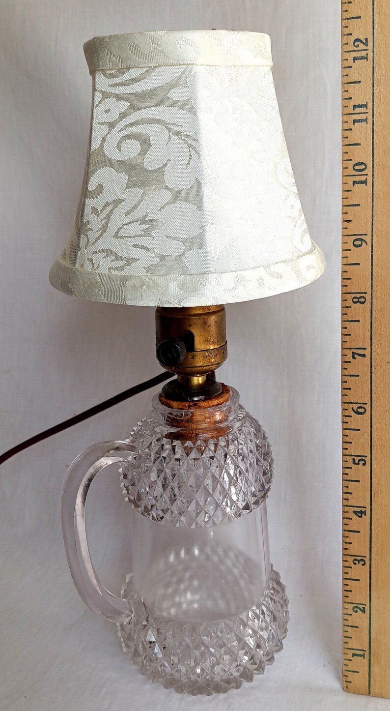May include: A vintage glass lamp base with a clear, faceted design and a handle. The lamp has a white fabric lampshade with a floral pattern. The lamp is approximately 20 cm tall.