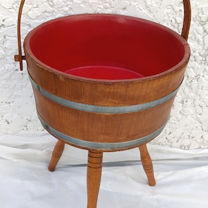 May include: A wooden bucket with a red interior and a handle. The bucket is on three wooden legs.