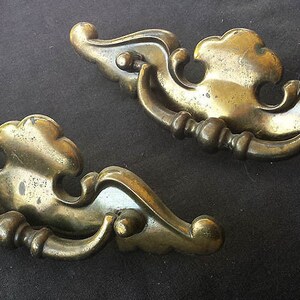 Op de afbeelding: Twee sierlijke messing ladegrepen met een decoratief, rolvormig ontwerp. De grepen hebben een vintage uitstraling en zijn in goede staat.