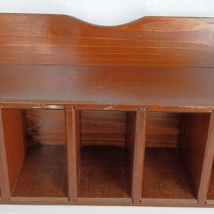May include: A brown wooden storage unit featuring a flat top surface with a curved back panel and five vertical compartments arranged in a row. The unit has a smooth, polished finish showing the natural wood grain throughout the structure.