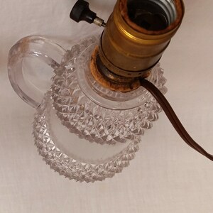 May include: A clear glass lamp base with a diamond-shaped pattern. The lamp base has a handle and a brass socket with a black switch.