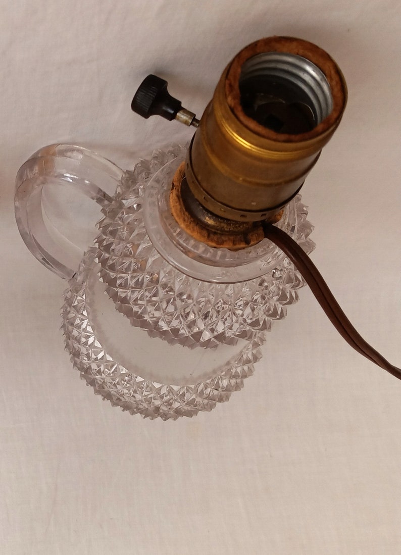 May include: A clear glass lamp base with a diamond-shaped pattern. The lamp base has a handle and a brass socket with a black switch.