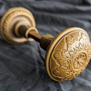 Puede incluir: Una perilla de puerta dorada con un diseño ornamentado y detallado. La perilla tiene una forma redonda y plana con un centro circular elevado. El diseño presenta intrincados patrones florales y remolinos.