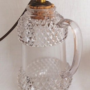 May include: A clear glass lamp with a diamond-shaped pattern and a handle. The lamp has a brass socket and a brown cord with a plug.