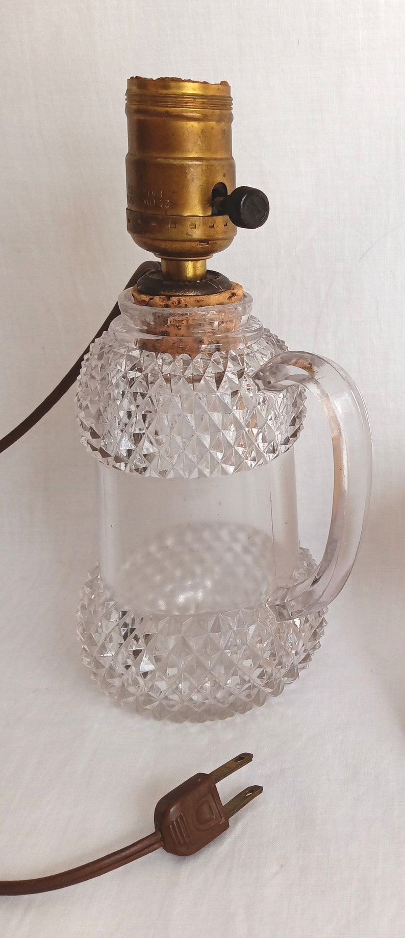 May include: A clear glass lamp with a diamond-shaped pattern and a handle. The lamp has a brass socket and a brown cord with a plug.