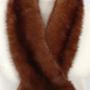 May include: A luxurious brown mink fur scarf draped over a white mannequin. The scarf is a rich, warm brown color with a soft, textured appearance. The fur appears dense and plush, suggesting high quality and warmth.