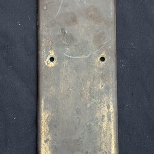 May include: A rectangular, antique brass door plate with six screw holes and six brass screws. The plate has a weathered appearance with signs of age and use, showing a mix of brown and gold tones.