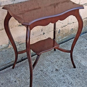 May include: A dark brown wooden side table with a polished rectangular top featuring curved edges and a scalloped apron. The table stands on four slender, curved legs that connect to a smaller, lower shelf positioned between the legs.
