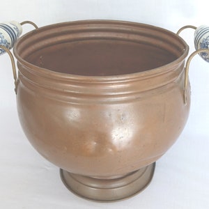 May include: A large copper planter with two porcelain handles. The handles are decorated with a blue and white floral pattern. The planter has a round base and a wide, round opening.