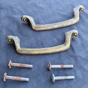 May include: Two antique brass drawer pulls with a scrolled design, and four screws. The pulls have a curved shape with decorative ends. The screws are a mix of sizes and finishes, some showing signs of age.
