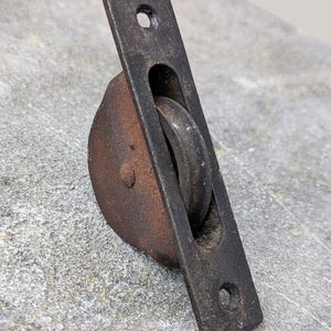May include: A vintage, rusty metal pulley with a rectangular mounting plate and a circular wheel. The plate has two holes for mounting. The pulley is likely for a door or window. The metal has a weathered, aged appearance.
