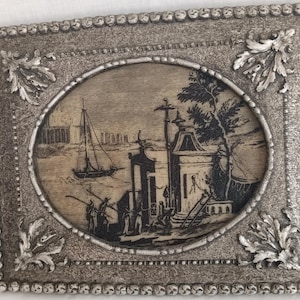 May include: A rectangular, silver-toned picture frame with an oval, sepia-toned image of a harbor scene. The frame has a beaded border and decorative corner accents. The image depicts a sailboat, buildings, and figures.