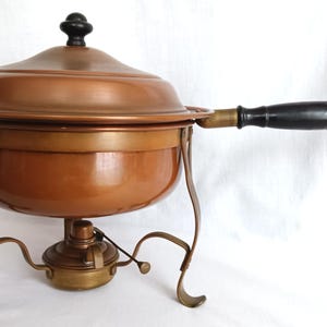 May include: A copper-colored chafing dish with a lid and a black handle. The dish sits on a brass-colored stand with curved legs and a burner below. The lid has a black knob.