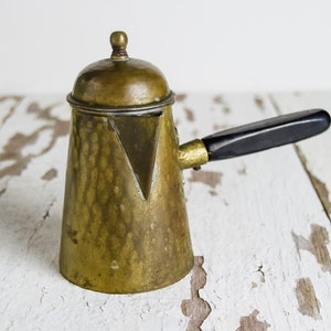 Puede incluir: Una cafetera de cobre con un mango negro y una tapa. La cafetera tiene un diseño triangular en la parte delantera y está sobre una superficie de madera blanca.