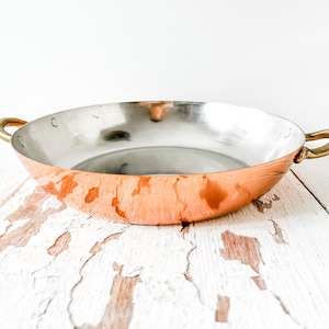 May include: A copper pan with brass handles. The pan has a shiny, silver interior and a copper exterior. The pan is sitting on a white, distressed wooden surface.
