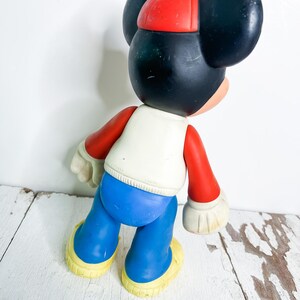 May include: A vintage Mickey Mouse figurine, featuring a red cap, black ears, and a white vest. The figure has red arms, blue pants, and yellow shoes. The toy is posed with its back to the viewer.