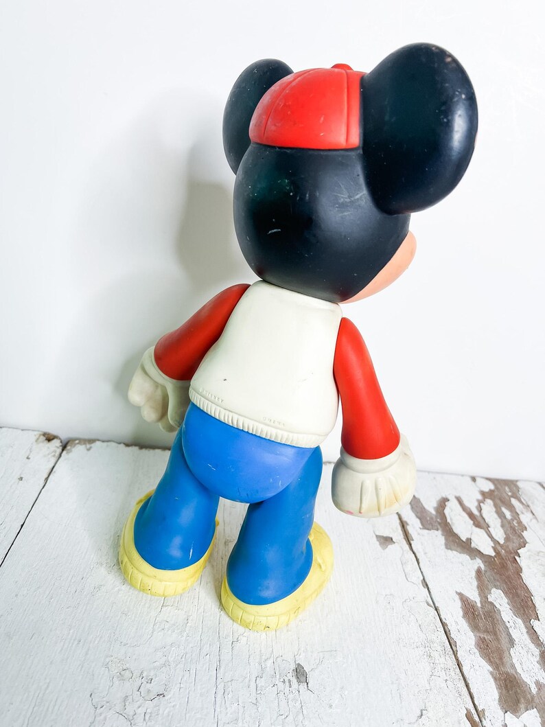 May include: A vintage Mickey Mouse figurine, featuring a red cap, black ears, and a white vest. The figure has red arms, blue pants, and yellow shoes. The toy is posed with its back to the viewer.