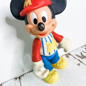 May include: A vintage Mickey Mouse figurine. The figure wears a red baseball cap with a yellow "M", a red and white striped shirt, blue pants, and yellow shoes. The figure has black ears, white gloves, and a black nose.