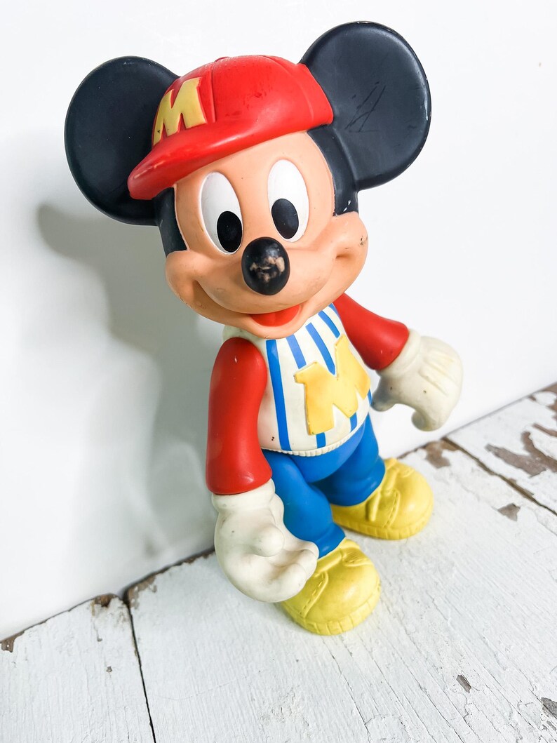 May include: A vintage Mickey Mouse figurine. The figure wears a red baseball cap with a yellow "M", a red and white striped shirt, blue pants, and yellow shoes. The figure has black ears, white gloves, and a black nose.