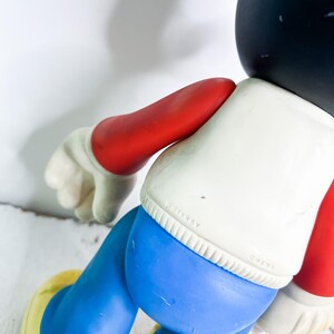 May include: A vintage Mickey Mouse figurine, featuring a black head, white shirt, blue shorts, red arms, and yellow shoes. The figurine has a classic design and is a collectible item.