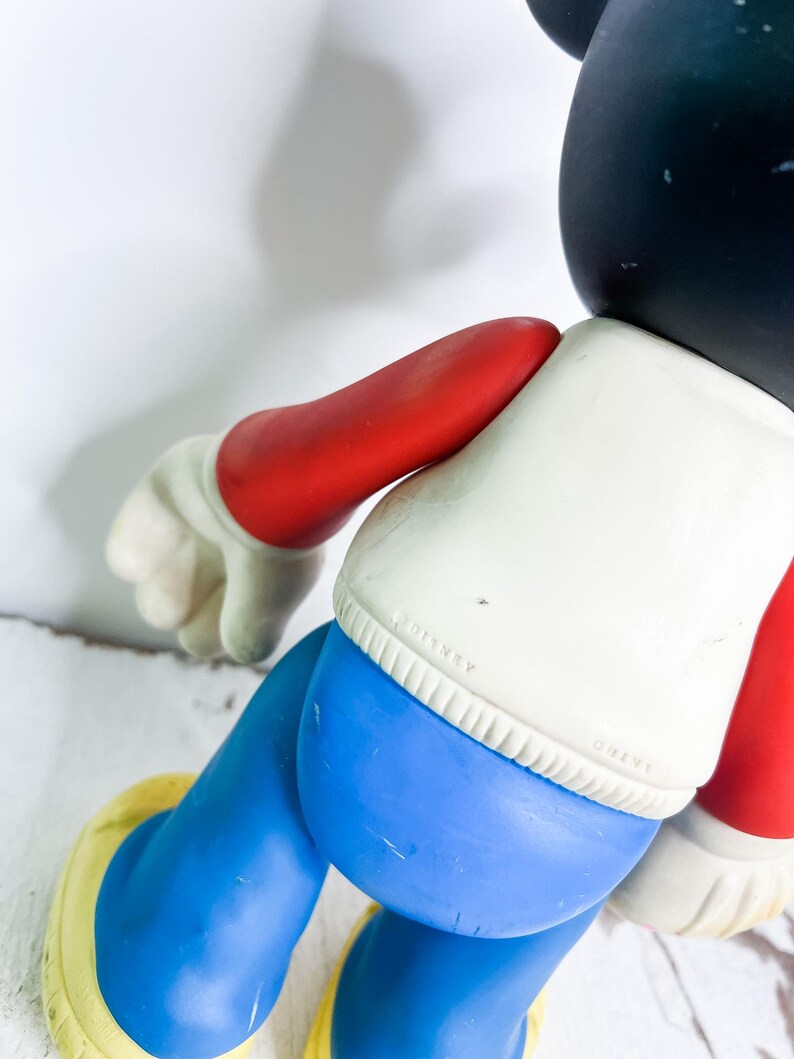 May include: A vintage Mickey Mouse figurine, featuring a black head, white shirt, blue shorts, red arms, and yellow shoes. The figurine has a classic design and is a collectible item.
