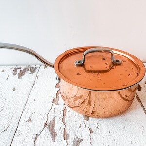 May include: A copper saucepan with a lid and a long, curved stainless steel handle. The pot has a shiny, reflective surface and sits on a distressed white wooden surface. The lid has a small, arched handle.