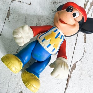 May include: Vintage Mickey Mouse toy figure. The figure is wearing a red hat, red long-sleeve shirt, blue pants, and yellow shoes. The shirt has a blue and white striped design with a yellow "M" on the front. The figure has white gloves and a smiling face.