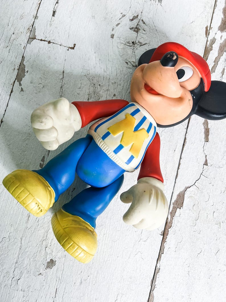 May include: Vintage Mickey Mouse toy figure. The figure is wearing a red hat, red long-sleeve shirt, blue pants, and yellow shoes. The shirt has a blue and white striped design with a yellow "M" on the front. The figure has white gloves and a smiling face.