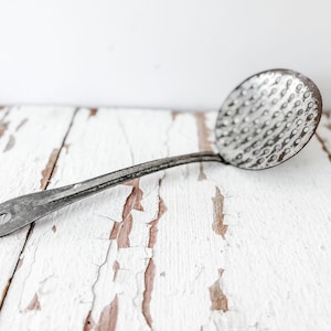May include: An antique metal skimmer with a long handle and a perforated, round head. The skimmer is made of a dark, aged metal and has a hole at the end of the handle for hanging. It rests on a distressed, white-painted wooden surface.