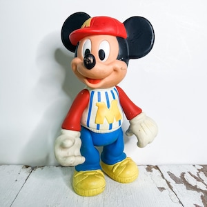 May include: A vintage Mickey Mouse figurine wearing a red cap, red and white striped shirt with a yellow "M", blue pants, yellow shoes, and white gloves. The classic cartoon character is standing against a white background.