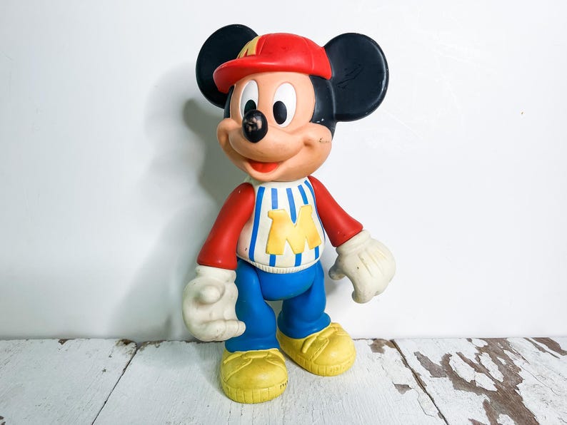 May include: A vintage Mickey Mouse figurine wearing a red cap, red and white striped shirt with a yellow "M", blue pants, yellow shoes, and white gloves. The classic cartoon character is standing against a white background.
