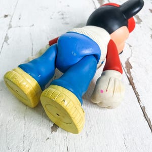 May include: A vintage Mickey Mouse figurine, lying on its stomach. The figure has a black head with red ears and a red hat. It wears a white sweater, blue pants, red arms, and yellow shoes.