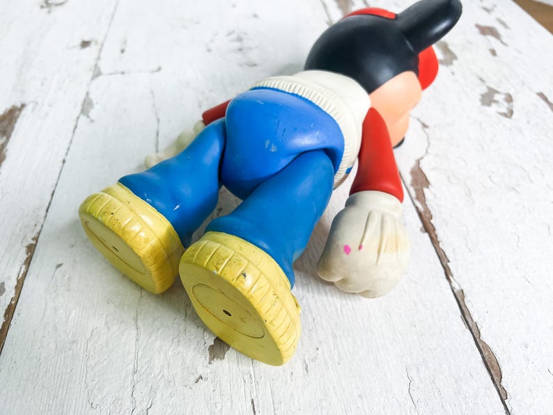 May include: A vintage Mickey Mouse figurine, lying on its stomach. The figure has a black head with red ears and a red hat. It wears a white sweater, blue pants, red arms, and yellow shoes.