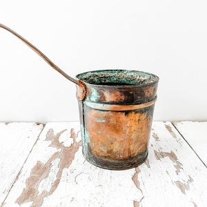 May include: A vintage copper ladle with a long handle. The ladle has a patina of age and is slightly tarnished. It is sitting on a white wooden surface.