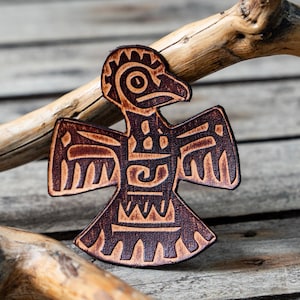 Może przedstawiać: Wycięty z brązowej skóry stylizowany ptak z misternymi geometrycznymi wzorami. Ptak ma dużą głowę i rozpostarte skrzydła.