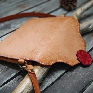 May include: A light brown leather crossbody bag with a red leather accent. The bag has a flap closure and a strap that can be adjusted.