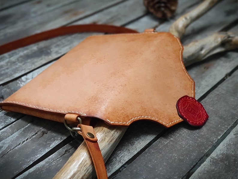 May include: A light brown leather crossbody bag with a red leather accent. The bag has a flap closure and a strap that can be adjusted.