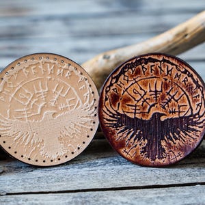 Puede incluir: Dos posavasos redondos de cuero con un diseño de un pájaro con las alas extendidas y runas alrededor del perímetro. Un posavasos es de color marrón claro y el otro es de color marrón oscuro.