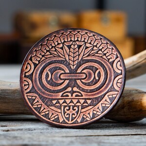 May include: A round, brown leather coaster with a detailed, carved design of a Polynesian tribal mask. The mask features a central eye with a swirling pattern and a geometric design around the edges.