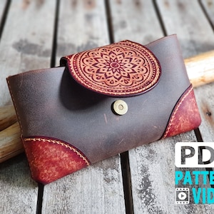 Può includere: Un portafoglio in pelle marrone con una patta in pelle rossa e un design decorativo a mandala. La patta ha una chiusura a bottone in ottone. Il portafoglio è su una superficie in legno.
